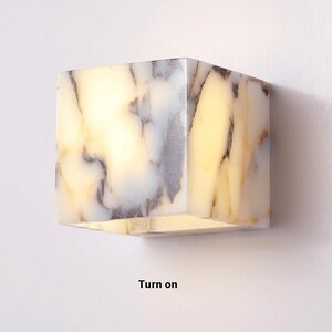 May include: A white marble cube wall sconce with a light inside. The light is turned on, illuminating the marble's natural veining. The sconce is mounted on a white wall. "Turn on" is written below the sconce.