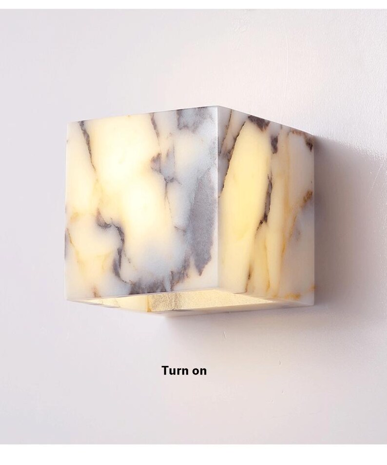 May include: A white marble cube wall sconce with a light inside. The light is turned on, illuminating the marble's natural veining. The sconce is mounted on a white wall. "Turn on" is written below the sconce.