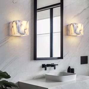 May include: A modern bathroom with a white marble countertop and sink. Two white marble cube-shaped wall sconces with warm light illuminate the room. A black framed mirror hangs above the sink.