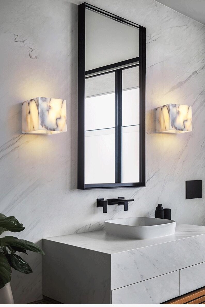 May include: A modern bathroom with a white marble countertop and sink. Two white marble cube-shaped wall sconces with warm light illuminate the room. A black framed mirror hangs above the sink.