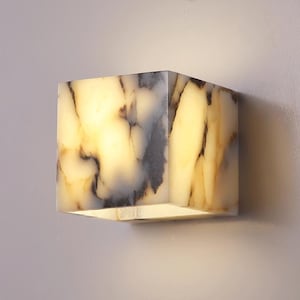 May include: A square-shaped wall sconce made of white alabaster with brown and grey veining. The sconce is lit and emits a warm glow.