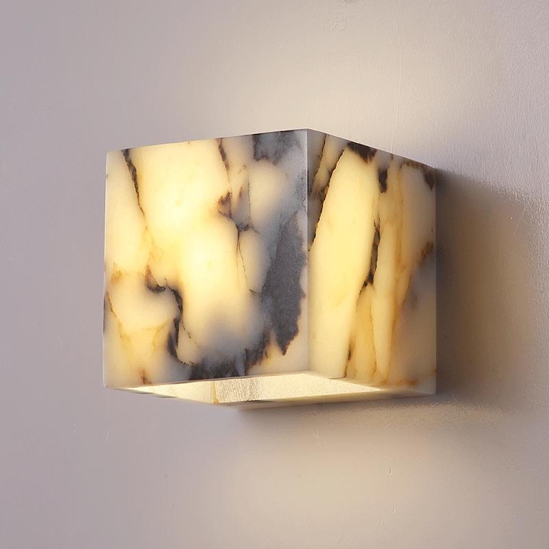 May include: A square-shaped wall sconce made of white alabaster with brown and gray veining. The sconce is lit and emits a warm glow.