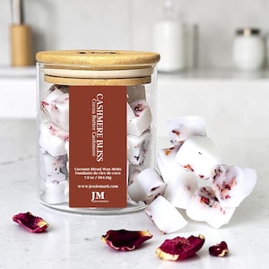 May include: A glass jar with a wooden lid containing white wax melts with red petal inclusions. The jar has a brown label with the text "Cashmere Bliss" and "Cocoa Butter Cashmere". Some melts are scattered around the jar.