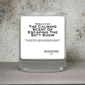 May include: A clear glass candle jar with a white label. The label reads "SMELLS LIKE THE CALMING SCENT OF ESCAPING THE SH*T SHOW." The candle is 255 grams and made of 100% soy wax, hand-poured in Toronto.