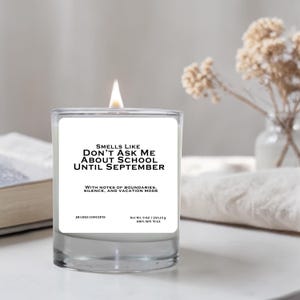 May include: A lit, clear glass candle with a white label that reads "Smells Like Don't Ask Me About School Until September." The candle has a 255.15 g net weight and is made of 100% soy wax. The candle is on a white surface.