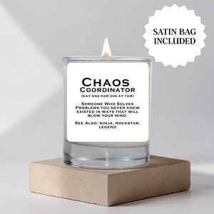 May include: A clear glass candle with a white label featuring the black text Chaos Coordinator, a phonetic pronunciation, and a humorous definition. The candle has a lit wick and is accompanied by a star-shaped graphic stating Satin Bag Included.