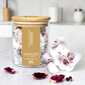May include: A glass jar with a wooden lid containing white wax melts with red petal inclusions. The label reads "KINDLE Honey Fig + Ginger" and "Coconut Blend Wax Melts 204.12g". Several melts are scattered around the jar on a white surface.