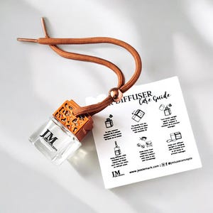 Peut inclure: Une petite bouteille en verre avec un bouchon en métal doré et un cordon brun. La bouteille est remplie d'un liquide clair et porte l'inscription "JM Luxe Concepts" sur l'étiquette. La bouteille est suspendue au cordon, qui est attaché à une carte portant l'inscription "Guide d'entretien du diffuseur de voiture".