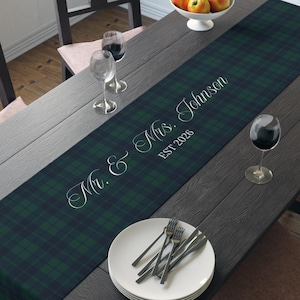 May include: A dark green and navy blue plaid table runner with the text "Mr. & Mrs. Johnson EST 2026" in white script. The runner is placed on a dark wooden table set for a meal, with plates, silverware, and wine glasses.