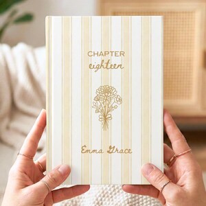 Pode incluir: Um livro de capa dura com uma capa listrada creme e bege. A capa apresenta o texto "CHAPTER eighteen" e "Emma Grace" em escrita dourada, juntamente com uma ilustração floral dourada.