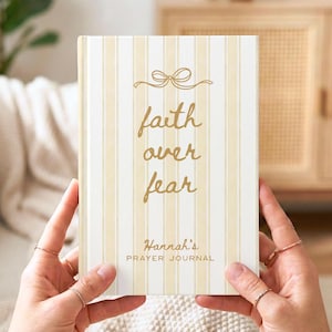 Puede incluir: Un diario crema con rayas verticales y letras doradas que dice "faith over fear". El diario está etiquetado como "Hannah's Prayer Journal" y presenta un diseño de lazo. El diario es sostenido por una persona.