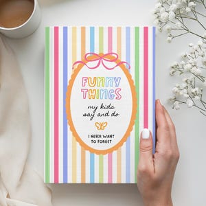 Op de afbeelding: Een kleurrijk notitieboek met verticale strepen in pasteltinten van roze, groen, geel en blauw. De kaft bevat de tekst "FUNNY THINGS my kids say and do I NEVER WANT TO FORGET" in een decoratief kader.