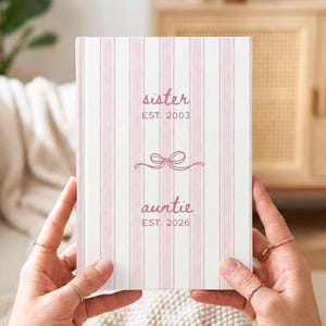 May include: A pink and white striped journal with the words "sister" and "auntie" in a script font. The journal has a bow graphic between the words. The journal is held by a person.