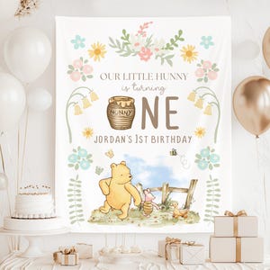 Puede incluir: Un telón de fondo blanco con ilustraciones de Winnie the Pooh y diseños florales para una fiesta de primer cumpleaños. El texto dice "Our Little Hunny is turning ONE, Jordan's 1st Birthday." Globos, una tarta y regalos están colocados delante.