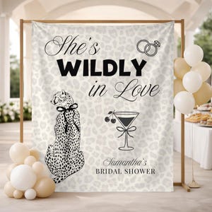 Op de afbeelding: Een achtergrond voor een bruidsdouche met een luipaardprint. De tekst "She's WILDLY in Love" wordt in het zwart weergegeven, samen met een illustratie van een cheetah, een cocktailafbeelding en de tekst "Samantha's BRIDAL SHOWER". Ballonnen in wit en beige staan links en rechts.
