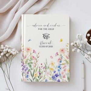 Puede incluir: Un libro de consejos y deseos de graduación con un diseño floral. La portada presenta el texto "advice and wishes FOR THE GRAD" y "Harrah CLASS OF 2026". El libro está decorado con flores de acuarela en rosa, morado y azul.