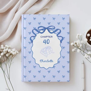 Könnte beinhalten: Ein hellblaues Buch mit Schleifenmuster. Das Cover zeigt ein dekoratives Oval mit dem Text "KAPITEL 40" und dem Namen "Charlotte". Das Design umfasst Illustrationen von Büchern und Schleifen.