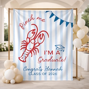 May include: A graduation backdrop with a red lobster illustration and the text "Pinch me, I'm a Graduate!" on a blue and white striped background. The backdrop also includes the text "Congrats Hannah CLASS OF 2026".