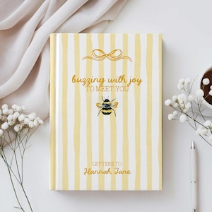Op de afbeelding: Een hardcover notitieboek met een geel en wit gestreepte kaft. De kaft heeft een gouden strik illustratie, een aquarel bij en de tekst "buzzing with joy to meet you" en "Letters to Hannah Jane". Een pen, bloemen en een kopje koffie staan op de achtergrond.