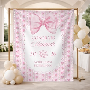 Könnte beinhalten: Ein rosa Abschluss-Hintergrund mit einer großen Schleife und dem Text "Congrats Hannah 2026 North Coast High School". Der Hintergrund hat ein Blumenmuster und wird von einer Holzkonstruktion umrahmt. Ballons in Weiß und Beige sind im Vordergrund.