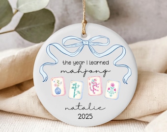 May include: A round white ceramic ornament featuring a blue line-art bow at the top. The text reads "the year I learned mahjong", "natalie", and "2025". Below the text are four mahjong tiles with illustrations of a vase, bamboo, dragon, and flower.