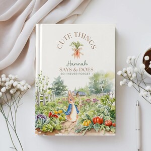May include: A hardcover journal with the text "CUTE THINGS" and "Hannah SAYS & DOES SO I NEVER FORGET." The cover features a watercolor illustration of a rabbit in a garden with pumpkins and vegetables. The journal is surrounded by flowers and a pen.