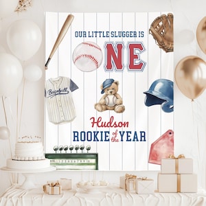 May include: A baseball-themed birthday banner featuring the text "Our Little Slugger is ONE" and "Hudson Rookie of the Year." The design includes a baseball, bat, glove, jersey, helmet, and a teddy bear holding a baseball.
