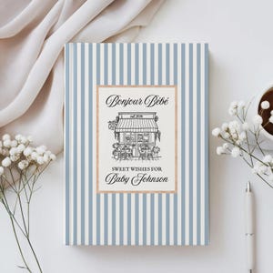 Può includere: Un libro degli ospiti per baby shower con una copertina a righe blu e bianche. Il libro presenta le parole "Bonjour Bébé" e "Sweet Wishes for Baby Johnson" in corsivo. Un'illustrazione dettagliata di un negozio è al centro.