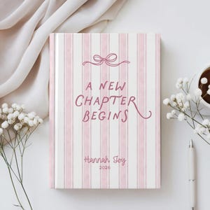 May include: A hardcover journal with a pink and white striped pattern. The cover features the text "A New Chapter Begins" in pink script, along with the name "Hannah Joy" and the year "2026". A bow graphic is at the top.
