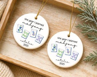May include: Two round white ceramic ornaments with gold hanging strings. Each features the text 'the year I learned mahjong', the name 'natalie', the year '2025', and a graphic of four mahjong tiles with blue, green, and pink illustrations.