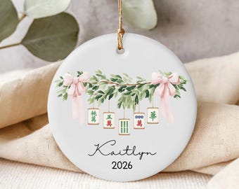 May include: A round white ceramic ornament featuring a watercolor illustration of green leafy branches adorned with two light pink bows. Five mahjong tiles hang from the branches. The name "Kaitlyn" and the year "2026" are printed in black text.