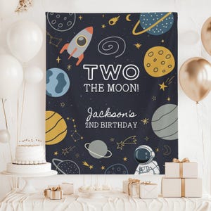 May include: A navy blue space-themed birthday banner with planets, stars, a rocket, and an astronaut. The banner reads "TWO THE MOON!" and "Jackson's 2nd Birthday". White and gold balloons, a cake, and wrapped gifts are also visible.