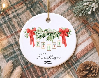 May include: A round white ceramic ornament featuring a watercolor illustration of green pine branches adorned with two red bows and five hanging mahjong tiles. The name Kaitlyn and the year 2025 are printed in black script and sans-serif font.