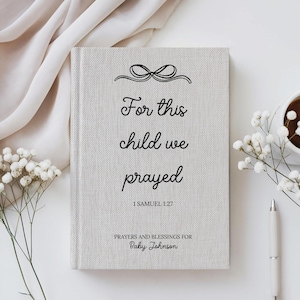 May include: A light grey linen-textured journal with the text "For this child we prayed" and "1 Samuel 1:27" in black script. The journal also includes the text "Prayers and Blessings for Baby Johnson". A pen, flowers, and a cup of coffee are in the background.