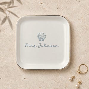 Może przedstawiać: Kwadratowa biała ceramiczna tacka ze złotą obwódką. Na środku znajduje się niebieska ilustracja muszli wykonana liniami oraz napis "Mrs. Johnson" niebieską kursywą. Tacka jest przeznaczona do przechowywania drobnych przedmiotów.