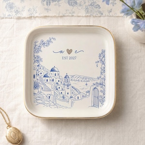 May include: A square white ceramic trinket dish with a gold-toned rim, featuring a blue ink illustration of a coastal village with domed buildings and stairs. The center displays the letters a and e separated by a heart, with the text EST 2027 below.