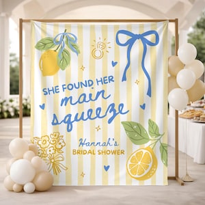 May include: A bridal shower backdrop with a yellow and white striped design. The backdrop features illustrations of lemons, a ring, and a blue bow. Text reads "She found her main squeeze" and "Hannah's Bridal Shower". Balloons are in the foreground.