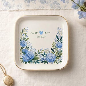 May include: A square white ceramic trinket dish with a gold-toned rim, featuring a watercolour illustration of blue hydrangea flowers and green leaves. The centre displays the lowercase letters a and e with a blue heart and the text EST 2027.