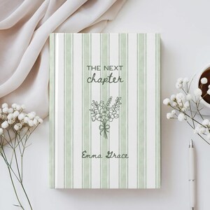 Puede incluir: Un diario de tapa dura con una cubierta a rayas verdes y blancas. El texto "THE NEXT chapter" y "Emma Grace" están impresos en verde, junto con una ilustración floral. Un bolígrafo y una taza de café están a la derecha.