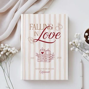May include: A hardback book with a beige and white vertical striped cover. The text reads Falling In Love, Est 2026, Autumn Nights, Future Mrs, Love Story, Notes For The Big Day, and Hannah. Graphics include two rings, a leaf, and a stack of books.