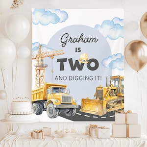 Puede incluir: Una pancarta de cumpleaños con el texto "¡Graham tiene dos años y le encanta!" presenta ilustraciones de vehículos de construcción, incluyendo un camión volquete y una excavadora, sobre un cielo azul con nubes. Globos blancos y dorados decoran el fondo.
