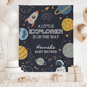 Może przedstawiać: Granatowe tło na baby shower z motywem kosmicznym. Projekt zawiera rakietę, planety, gwiazdy i tekst "A Little Explorer Is On The Way". Baby shower jest dla Hannah.