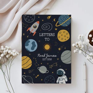Könnte beinhalten: Ein dunkelblaues Notizbuch mit Weltraum-Design. Es zeigt Illustrationen von Planeten, Sternen, einer Rakete und einem Astronauten. Auf dem Cover steht "LETTERS TO Noah James EST 2028".