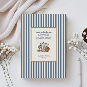 May include: A baby shower guest book with a blue and white striped cover. The center features a brown frame with the text "sweet wishes for our LITTLE SLUGGER" and a baseball-themed illustration. Includes a teddy bear, baseball jersey, and glove.