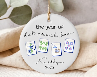 May include: A round white ceramic ornament hanging from a gold string. The text reads the year of dot crack bam, Kaitlyn, and 2025. Below the text are four mahjong tiles featuring a blue bird, green bamboo, a monkey with an umbrella, and five dots.