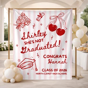 Puede incluir: Una pancarta de graduación a rayas rosas y blancas con ilustraciones rojas de flores, cerezas, un diploma y un birrete. El texto dice "Shirley She's Not Graduated! Congrats Hannah Class of 2026 North Coast High School."