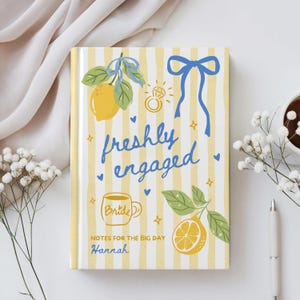 May include: A notebook with a yellow and white striped cover, featuring illustrations of lemons, a ring, and blue bows. The words "freshly engaged" are written in blue script. The notebook also includes the text "Notes for the Big Day" and a name.