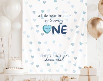 May include: A white birthday banner with blue heart patterns and the text "a little heartbreaker is turning ONE". The banner also includes the text "Happy Birthday Savannah". White and gold balloons, a cake, and wrapped gifts are also visible.