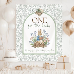 Könnte beinhalten: Ein dekoratives Schild mit floralem Hintergrund und einer Peter Hase-Illustration. Auf dem Schild steht "ONE for the books" und "Happy 1st Birthday Hayden". Das Bild zeigt Ballons, einen Kuchen und verpackte Geschenke.