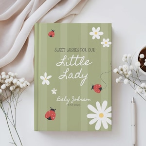 May include: A personalized baby journal with a green striped cover. The cover features the text "Sweet Wishes for Our Little Lady" and "Baby Johnson EST 2026". Adorned with ladybugs and white daisies.